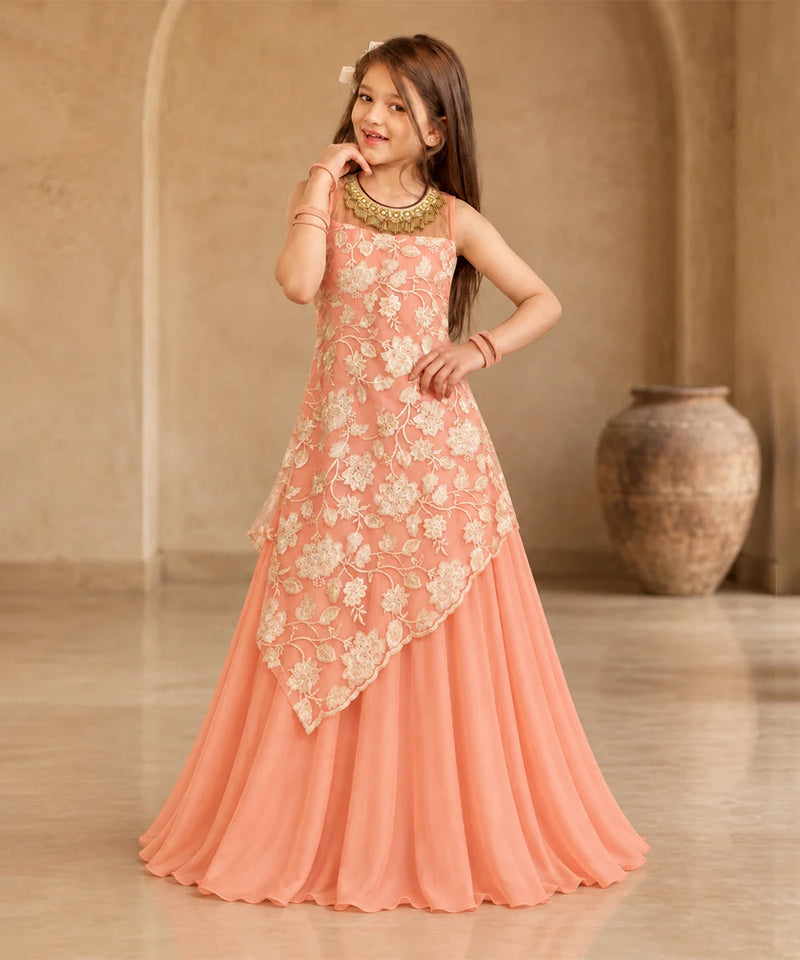 A young girl modeling a peach-colored sleeveless ethnic gown with cream floral embroidery and a draped asymmetrical overlay, set against a warm beige architectural background with a large clay pot.