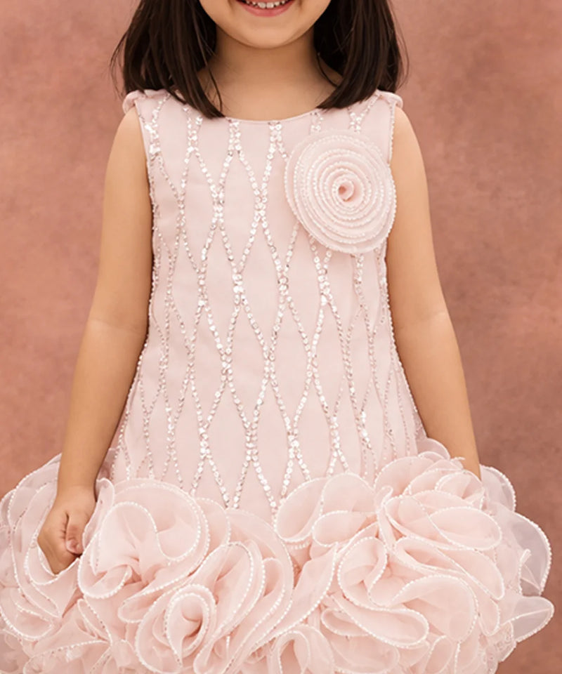 A young girl wearing a sleeveless light pink dress with white beaded geometric patterns, a large fabric rosette on the chest, and a voluminous ruffled tulle skirt, posed against a textured dusty pink background.