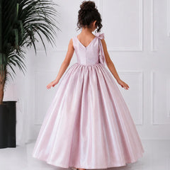 Young girl in a pink dress standing against a white wall with a plant to the left.