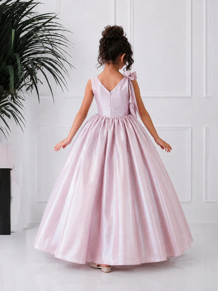 Young girl in a pink dress standing against a white wall with a plant to the left.