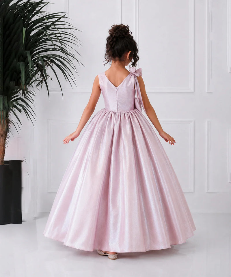 Young girl in a pink dress standing against a white wall with a plant to the left.