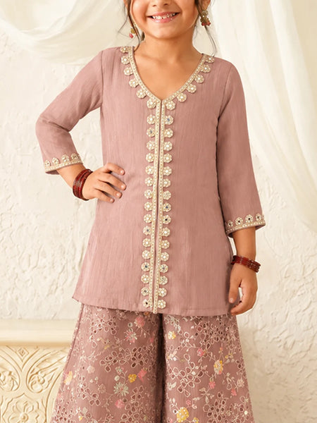 Child wearing a mauve embroidered outfit against a white curtain background