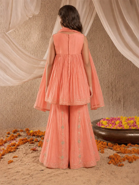 Woman in a peach traditional outfit standing in front of draped fabric with floral decorations on the ground.