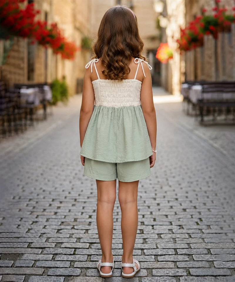 A young girl viewed from behind wearing a sage green two-piece set consisting of a sleeveless smocked top with tie straps and matching shorts, standing on a cobblestone street.