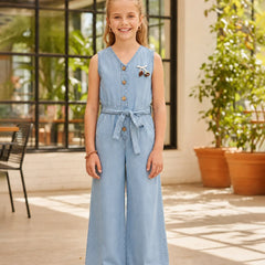 A young girl stands in a bright, modern indoor patio setting wearing a light blue denim jumpsuit. The sleeveless jumpsuit features a buttoned V-neck, a matching fabric tie belt at the waist, and wide legs.