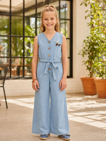 A young girl stands in a bright, modern indoor patio setting wearing a light blue denim jumpsuit. The sleeveless jumpsuit features a buttoned V-neck, a matching fabric tie belt at the waist, and wide legs.