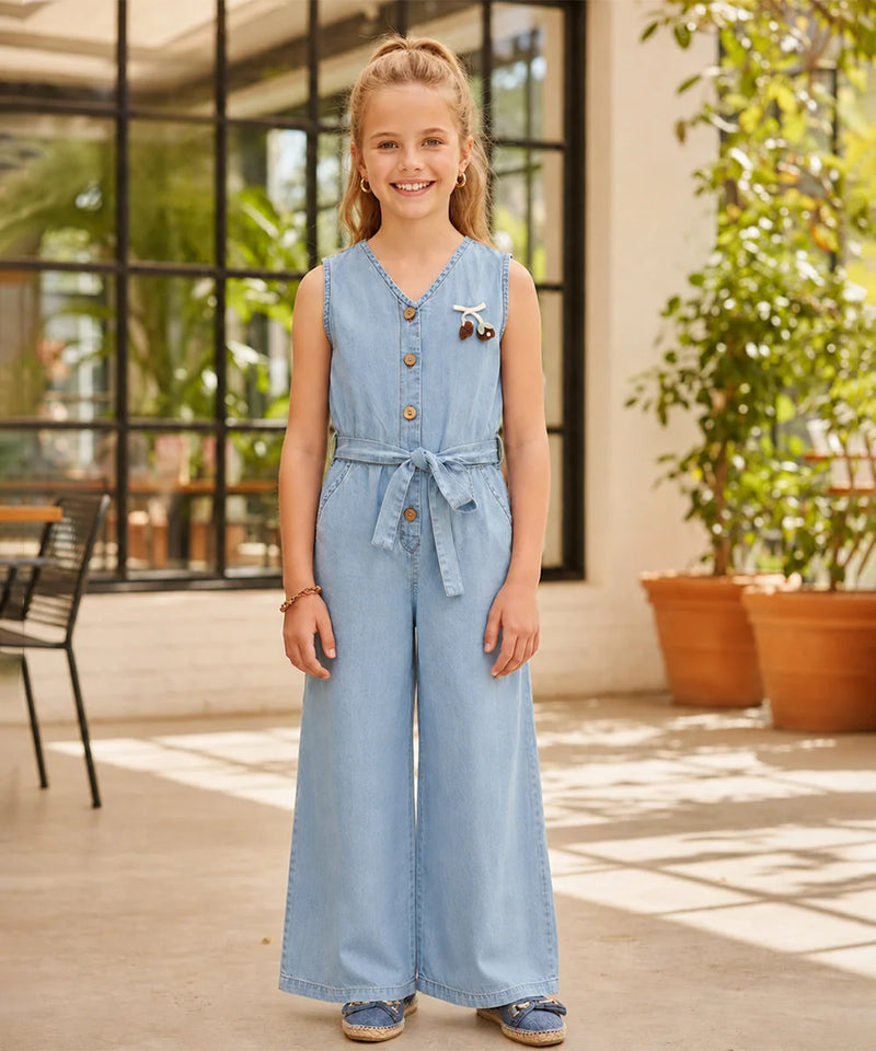 A young girl stands in a bright, modern indoor patio setting wearing a light blue denim jumpsuit. The sleeveless jumpsuit features a buttoned V-neck, a matching fabric tie belt at the waist, and wide legs.
