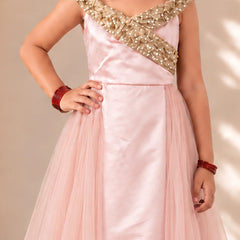 A girl wearing a light pink formal gown with a heavily embellished gold beaded bodice and a flowing tulle skirt, posing against a neutral beige background.