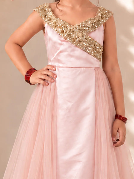 A girl wearing a light pink formal gown with a heavily embellished gold beaded bodice and a flowing tulle skirt, posing against a neutral beige background.