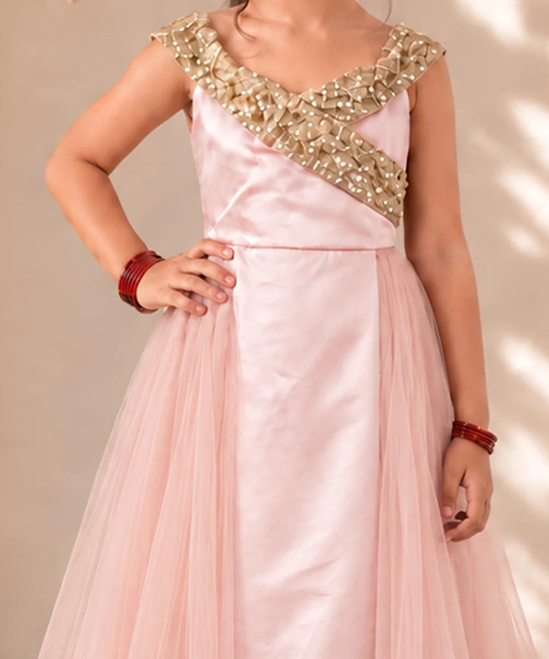 A girl wearing a light pink formal gown with a heavily embellished gold beaded bodice and a flowing tulle skirt, posing against a neutral beige background.