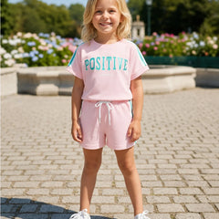 A young girl stands outdoors on a paved walkway wearing a light pink two-piece lounge set with a t-shirt that says 'POSITIVE' in teal and matching shorts with a white drawstring. She is wearing white sneakers, with a garden and fountain visible in the background.
