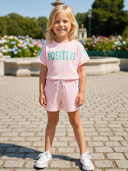A young girl stands outdoors on a paved walkway wearing a light pink two-piece lounge set with a t-shirt that says 'POSITIVE' in teal and matching shorts with a white drawstring. She is wearing white sneakers, with a garden and fountain visible in the background.