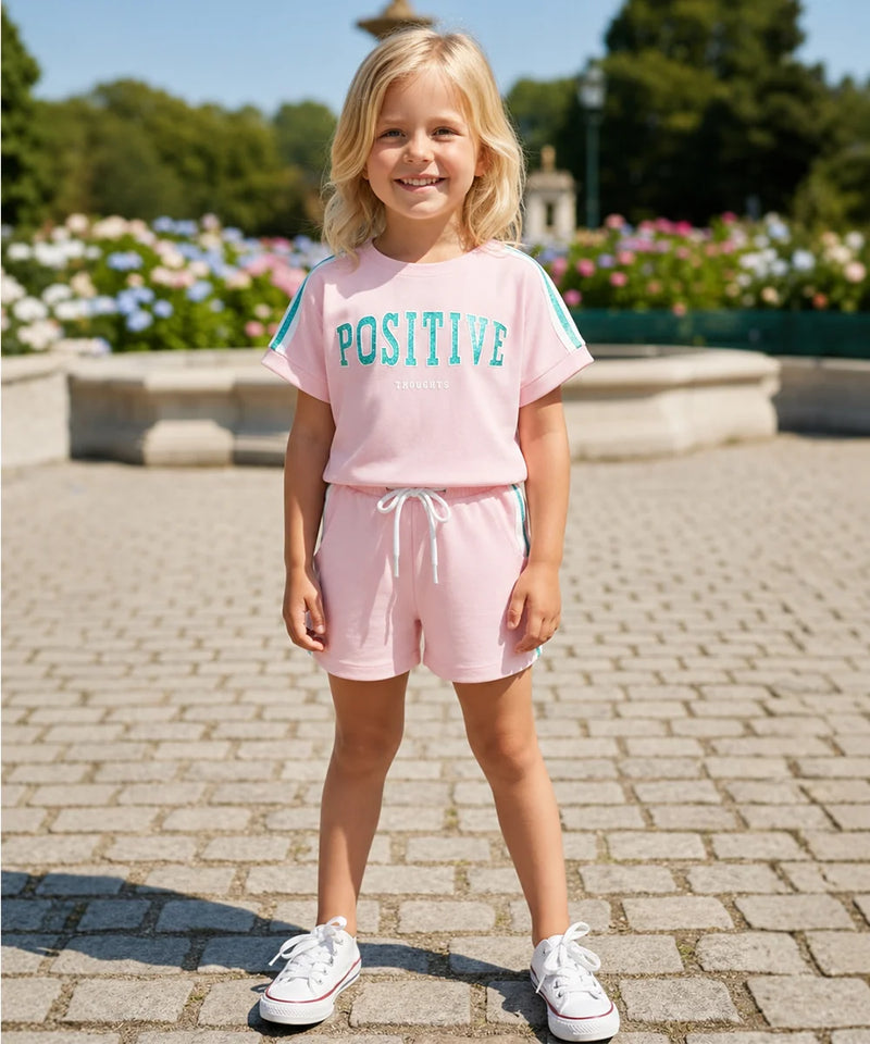 A young girl stands outdoors on a paved walkway wearing a light pink two-piece lounge set with a t-shirt that says 'POSITIVE' in teal and matching shorts with a white drawstring. She is wearing white sneakers, with a garden and fountain visible in the background.