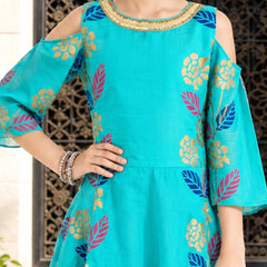 A woman wearing a turquoise cold-shoulder kurta with colorful floral and leaf patterns in blue, pink, and gold, paired with silver earrings.