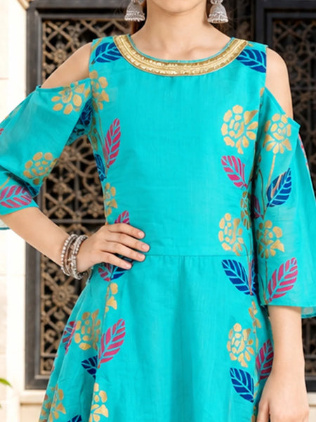 A woman wearing a turquoise cold-shoulder kurta with colorful floral and leaf patterns in blue, pink, and gold, paired with silver earrings.