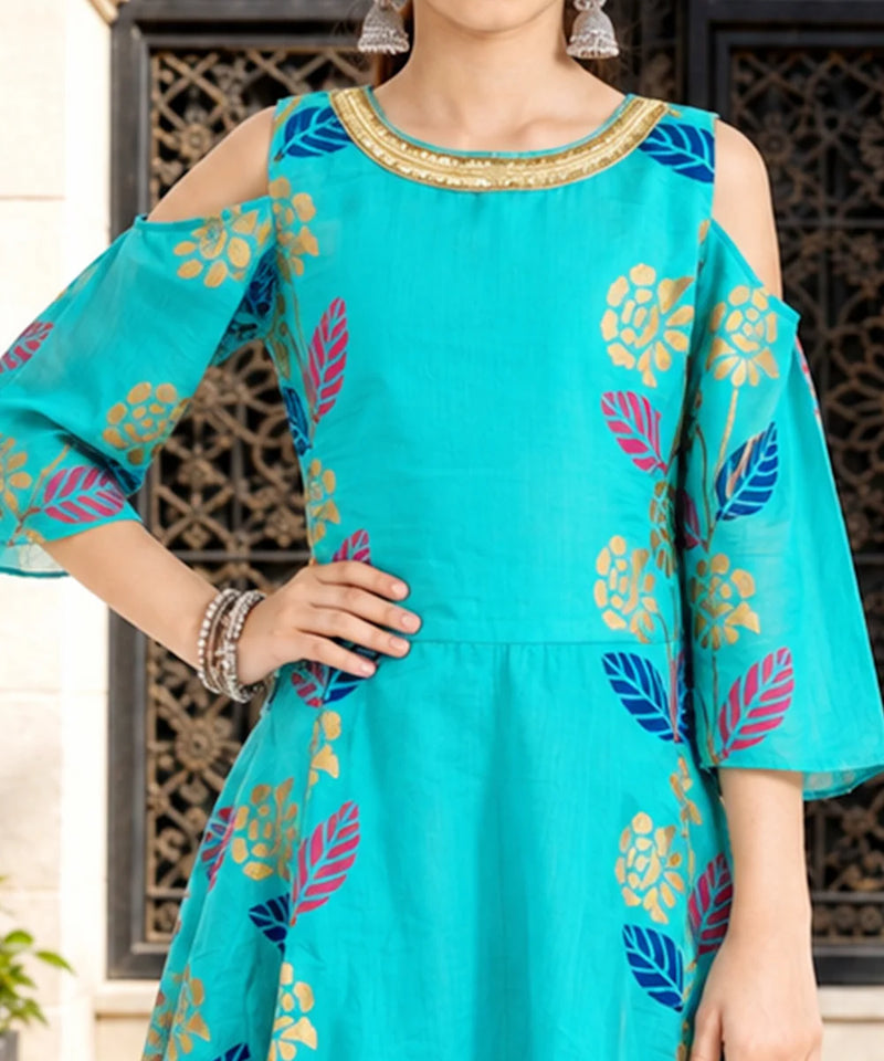 A woman wearing a turquoise cold-shoulder kurta with colorful floral and leaf patterns in blue, pink, and gold, paired with silver earrings.