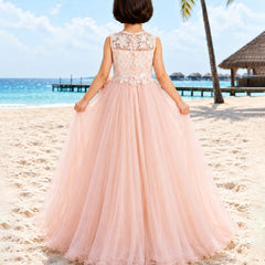 A young girl viewed from behind wearing a pale pink ball gown with a lace bodice and tulle skirt, standing on a sandy beach with the ocean and palm trees in the background.