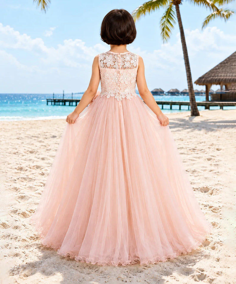 A young girl viewed from behind wearing a pale pink ball gown with a lace bodice and tulle skirt, standing on a sandy beach with the ocean and palm trees in the background.