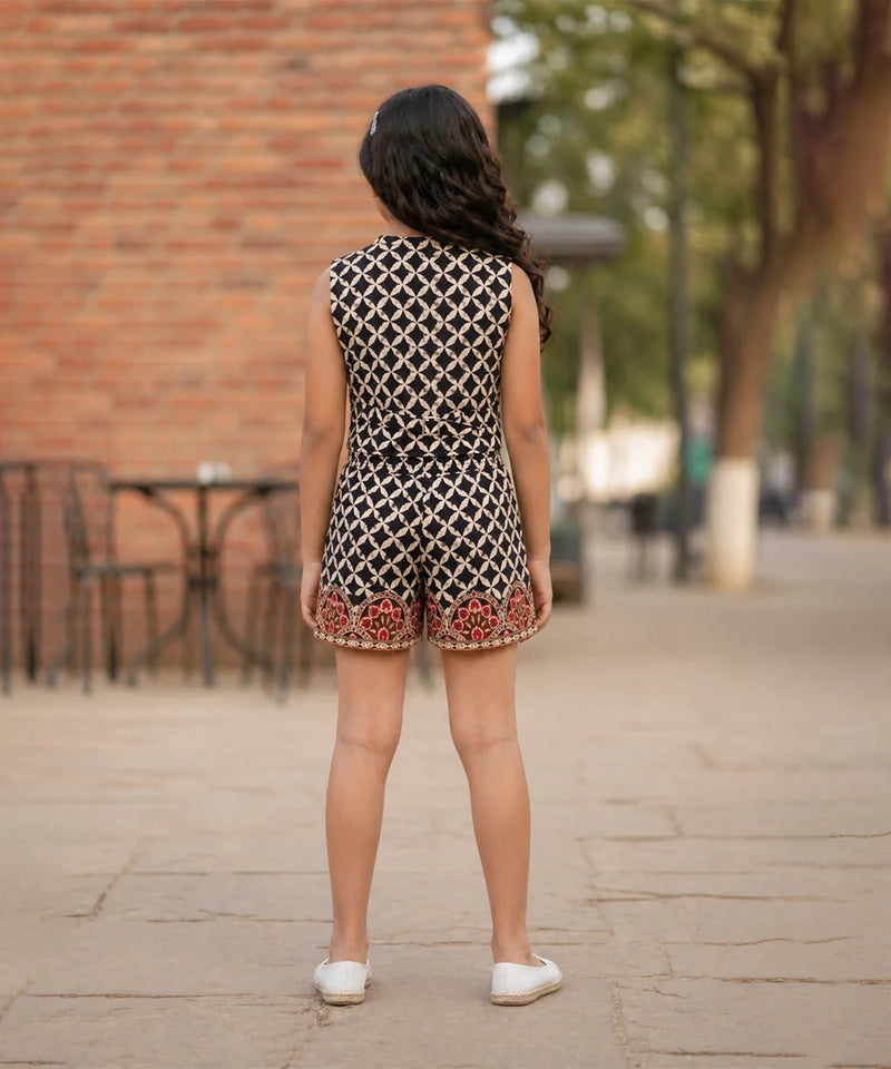 Person wearing a patterned romper standing on a paved area with a brick wall and trees in the background.