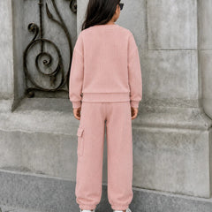 A child viewed from behind wearing a matching dusty pink sweatshirt and cargo pants set, standing outdoors against a gray stone wall.
