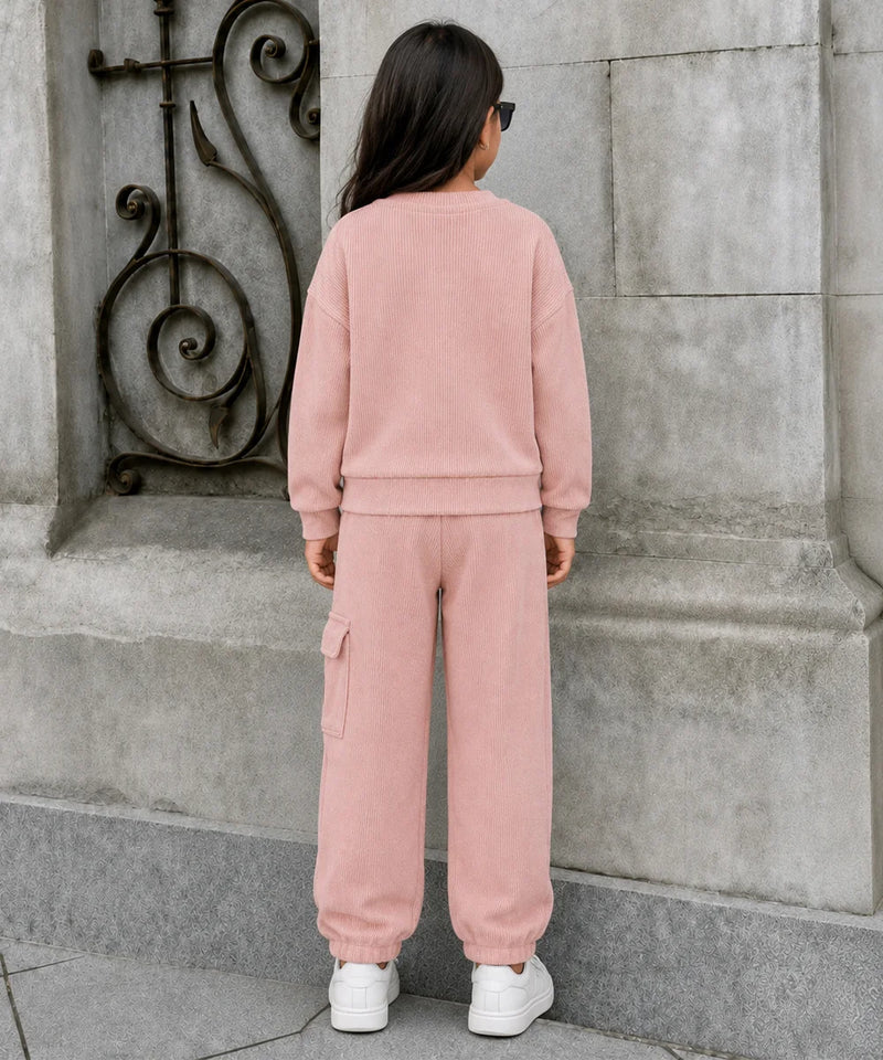 A child viewed from behind wearing a matching dusty pink sweatshirt and cargo pants set, standing outdoors against a gray stone wall.
