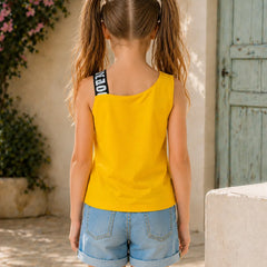 A young girl with blonde pigtails is shown from behind wearing a bright yellow sleeveless tank top with a black and white striped shoulder strap, paired with light blue denim shorts, set against a Mediterranean-style stone wall background.