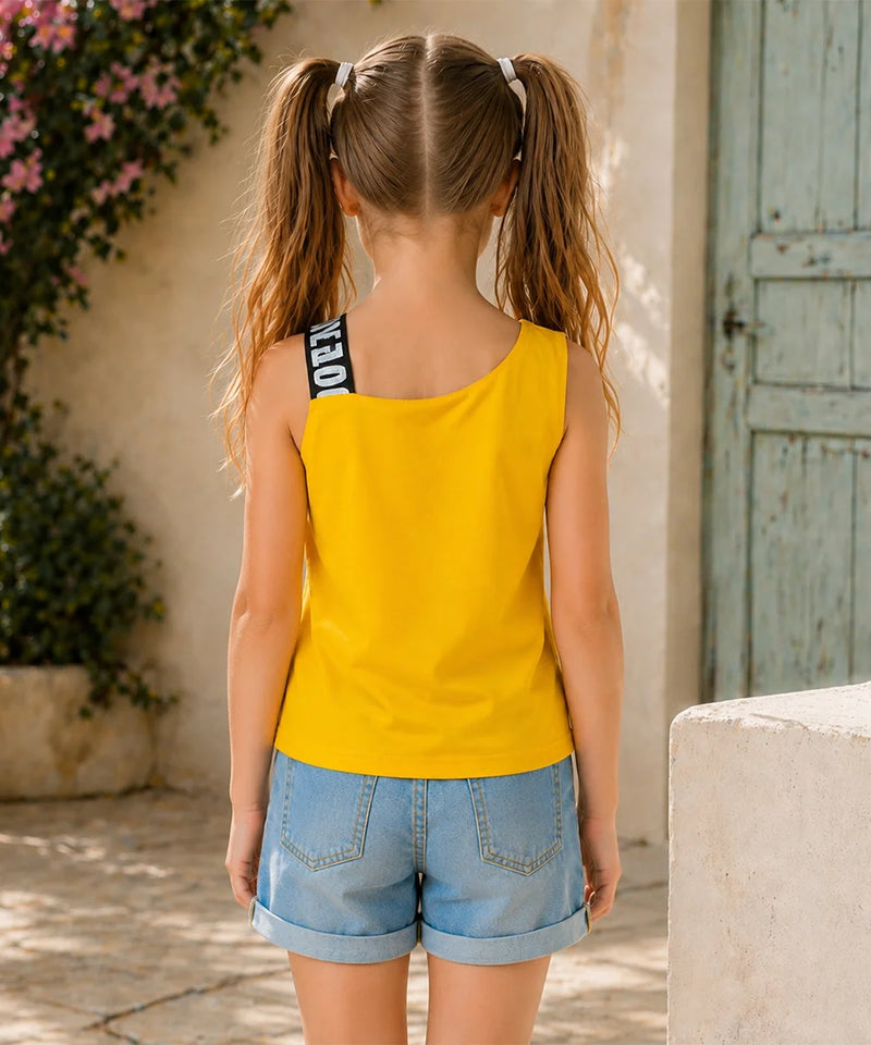 A young girl with blonde pigtails is shown from behind wearing a bright yellow sleeveless tank top with a black and white striped shoulder strap, paired with light blue denim shorts, set against a Mediterranean-style stone wall background.
