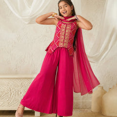 Young girl in a pink traditional outfit with gold patterns, standing against a white curtain.