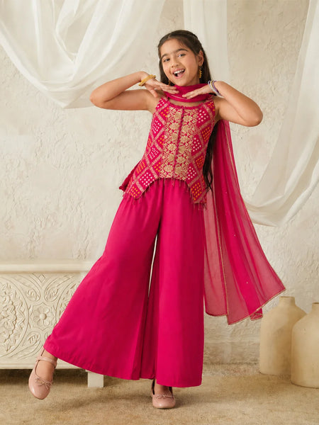 Young girl in a pink traditional outfit with gold patterns, standing against a white curtain.