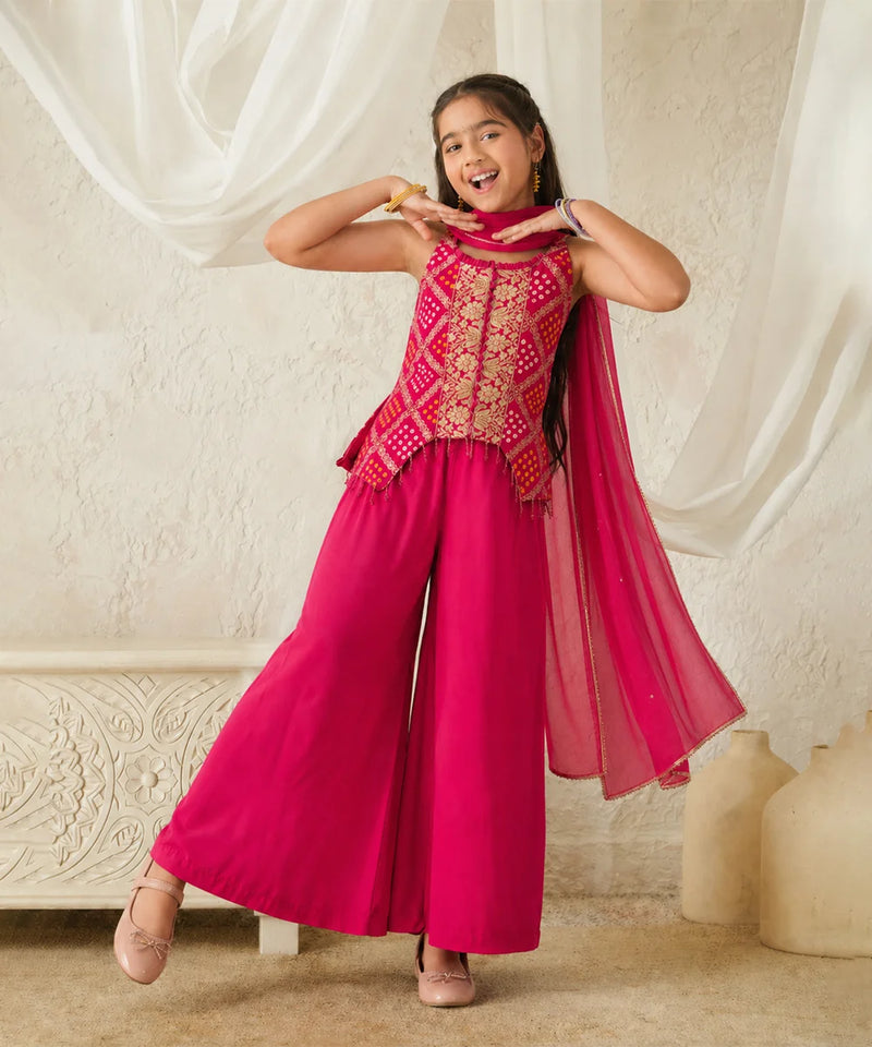 Young girl in a pink traditional outfit with gold patterns, standing against a white curtain.