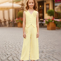 A young girl stands on a cobblestone street wearing a pale yellow sleeveless vest and matching wide-leg pants with gold floral embroidery, photographed outdoors in front of a blurred storefront.