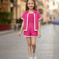 A young girl walking down a blurred city street wearing a bright pink two-piece outfit with white zigzag trim, consisting of a short-sleeve collared top and matching shorts.