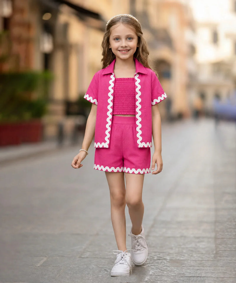 A young girl walking down a blurred city street wearing a bright pink two-piece outfit with white zigzag trim, consisting of a short-sleeve collared top and matching shorts.