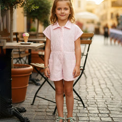 A young girl stands on a cobblestone street wearing a light pink short-sleeve eyelet romper with a collar, paired with green patterned sandals and a green bow in her hair.
