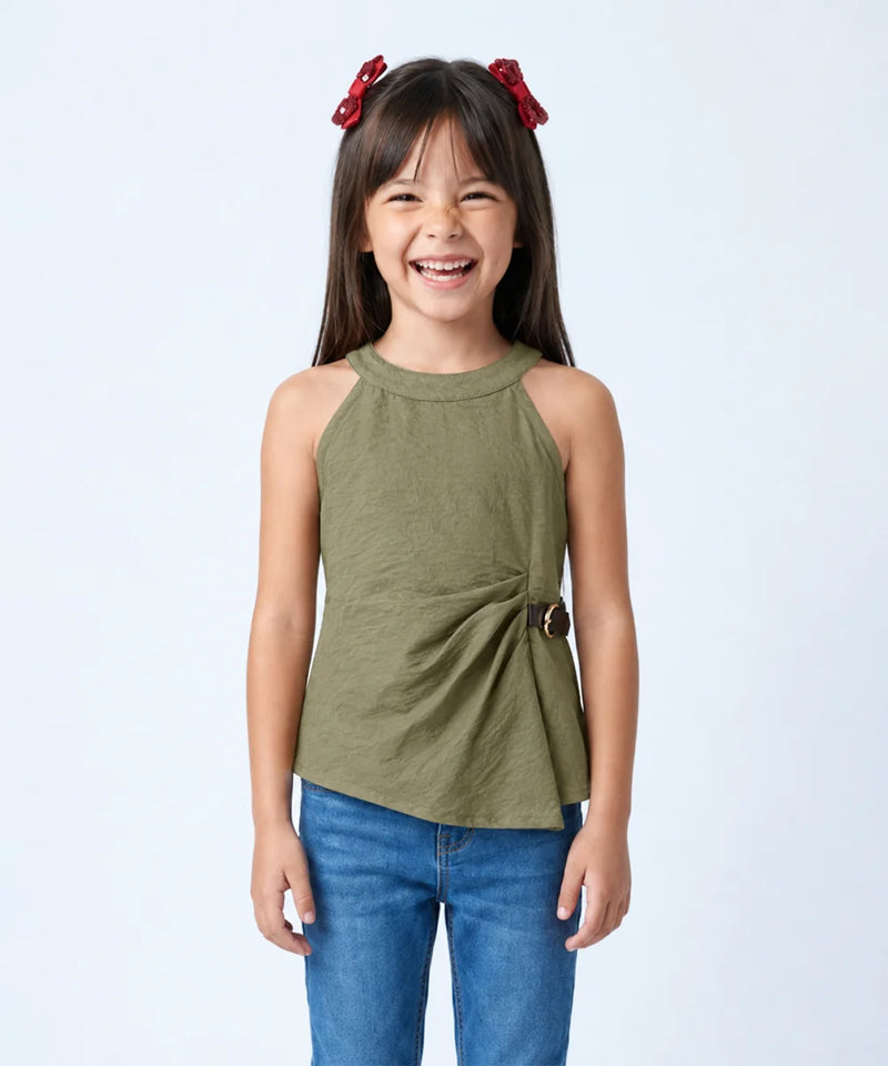 A young girl with dark hair and red bow clips is smiling, wearing an olive green sleeveless wrap-style top with blue jeans against a light gray studio background.