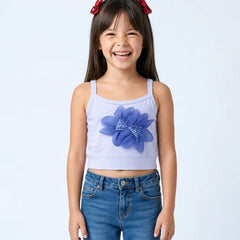 A young girl with dark hair and red bows in her hair is smiling, wearing a light lavender sleeveless crop top with a large blue flower applique on the front, paired with blue jeans, against a light gray studio background.
