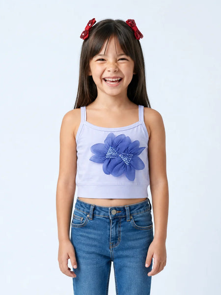 A young girl with dark hair and red bows in her hair is smiling, wearing a light lavender sleeveless crop top with a large blue flower applique on the front, paired with blue jeans, against a light gray studio background.