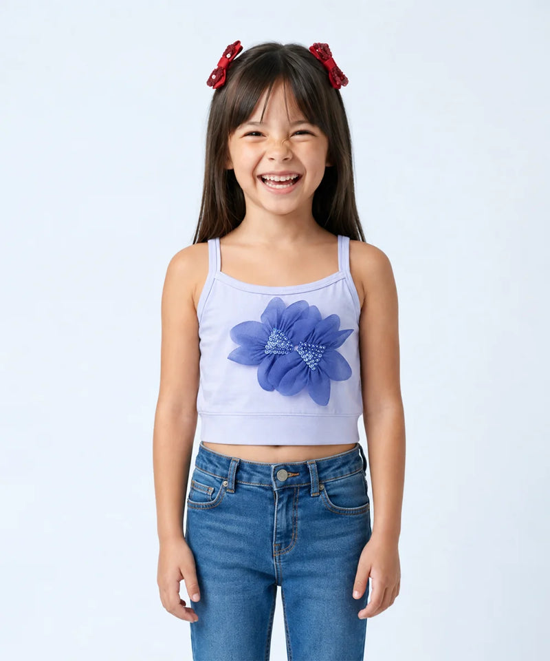 A young girl with dark hair and red bows in her hair is smiling, wearing a light lavender sleeveless crop top with a large blue flower applique on the front, paired with blue jeans, against a light gray studio background.
