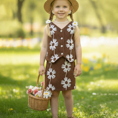 Young girl in a brown dress with white floral patterns, holding a basket of flowers, standing in a grassy field.
