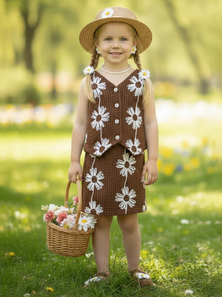 Young girl in a brown dress with white floral patterns, holding a basket of flowers, standing in a grassy field.