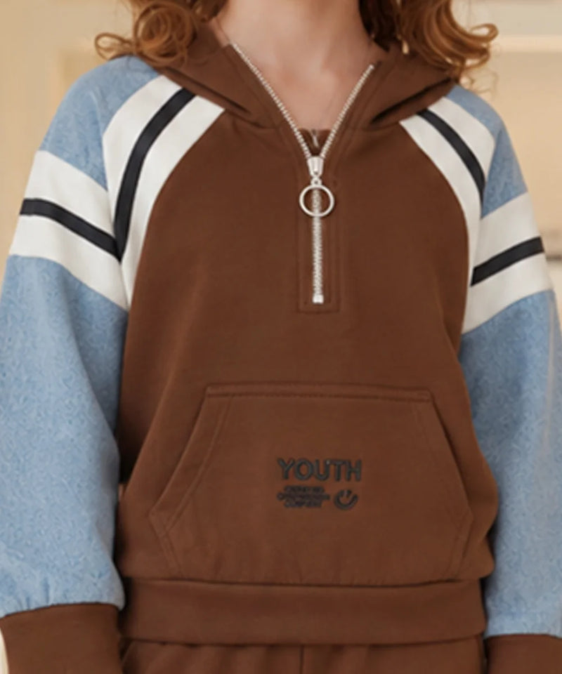 A person wearing a brown half-zip hoodie with light blue sleeves featuring white and black stripes, and a silver zipper pull with a ring detail. The front has a kangaroo pocket with 'YOUTH' embroidered on it.