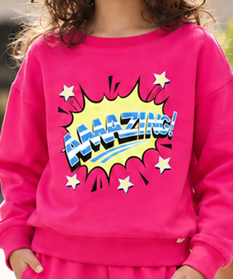 A child wearing a bright pink sweatshirt with a large comic-style 'AMAZING!' graphic in yellow, blue, and white on the front, paired with matching pink bottoms.