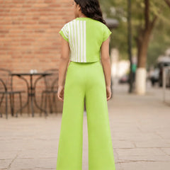 A woman viewed from behind wearing a bright lime green two-piece outfit consisting of a short-sleeve top with white vertical stripes and matching wide-leg pants, standing on a paved outdoor walkway.