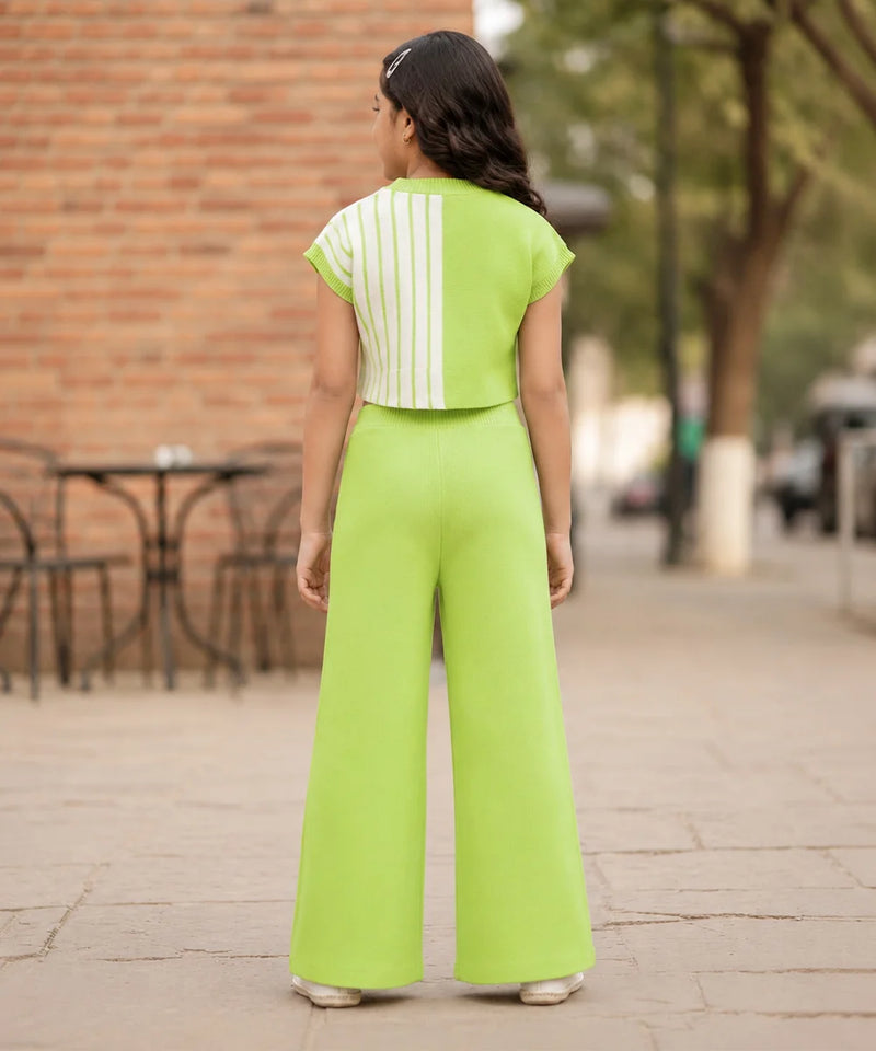 A woman viewed from behind wearing a bright lime green two-piece outfit consisting of a short-sleeve top with white vertical stripes and matching wide-leg pants, standing on a paved outdoor walkway.