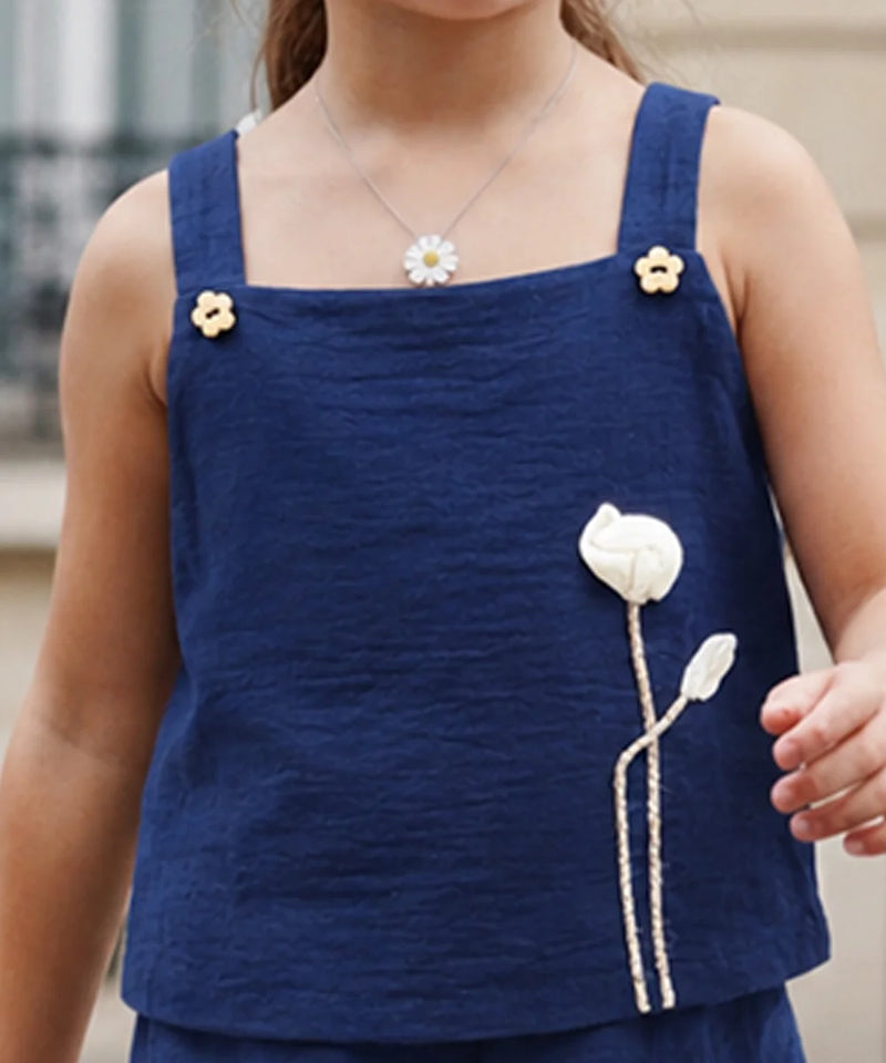 A close-up of a child wearing a navy blue sleeveless top with thick shoulder straps, small yellow flower embellishments, and a large white flower applique on the front.