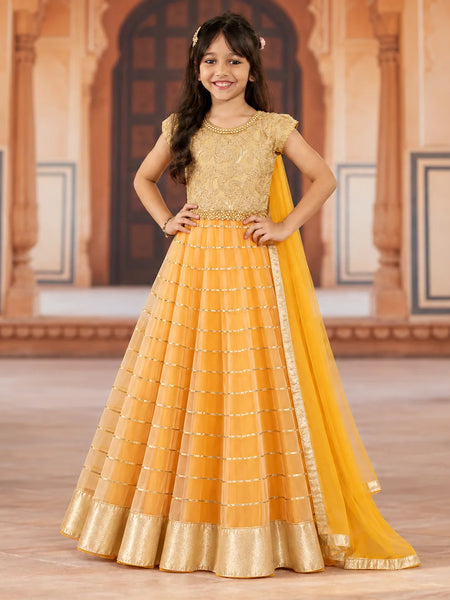 A young girl modeling a traditional Indian anarkali gown in mustard yellow and gold, standing in front of a warm-toned architectural background with arches.