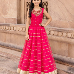 A young girl stands outdoors in front of a beige stone building, wearing a bright pink sleeveless Anarkali gown with gold horizontal stripes and a gold border hem.
