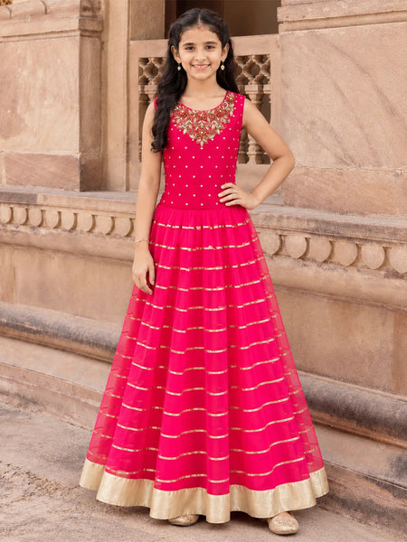 A young girl stands outdoors in front of a beige stone building, wearing a bright pink sleeveless Anarkali gown with gold horizontal stripes and a gold border hem.