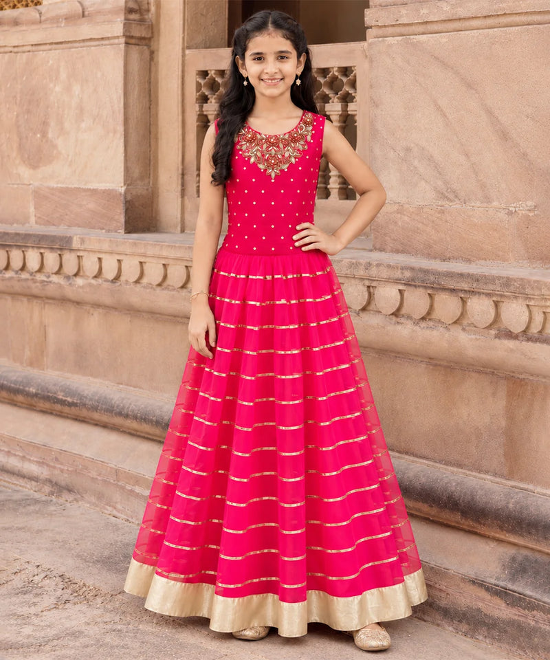 A young girl stands outdoors in front of a beige stone building, wearing a bright pink sleeveless Anarkali gown with gold horizontal stripes and a gold border hem.
