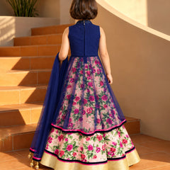 A young girl viewed from behind wearing a traditional Indian anarkali-style dress with a navy blue sleeveless bodice and a heavily embroidered pink floral skirt with a matching navy dupatta, standing on terracotta stairs.